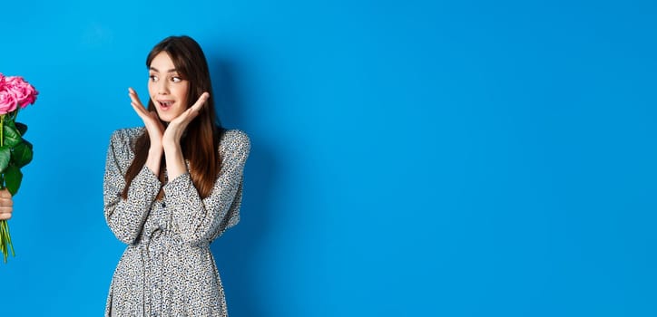 Valentines day concept. Romantic girl looking surprised at hand with flowers, woman receiving bouquet of pink roses, standing on blue background.