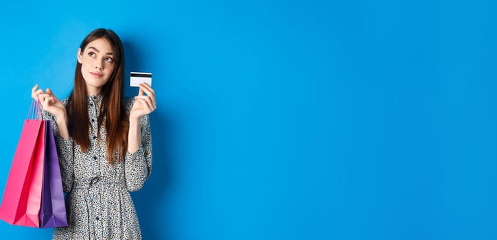 Dreamy girl looking at logo and showing plastic credit card, holding shopping bags, standing on blue background.