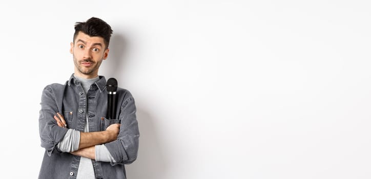 Image of funny male entertainer or singer, holding microphone in pocket of jacket and look at camera with arms crossed, standing against white background.