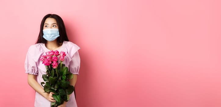 Romantic asian girl in medical mask looking aside at empty space with dreamy eyes, holding bouquet of roses for Valentines day, having date with lover during pandemic.