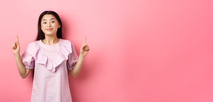 Beautiful young asian woman in romantic dress pointing fingers up, smiling and gladly showing advertisement, standing on pink background.