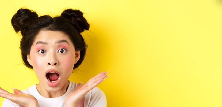 Close-up of excited asian woman with glamour makeup and hairstyle, screaming of amazement, open mouth and look at camera in awe, yellow background.