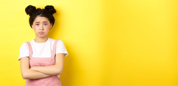 Sad asian teen girl sulking, cross arms on chest, pouting and frowning, complaining on unfair thing, standing against yellow background.