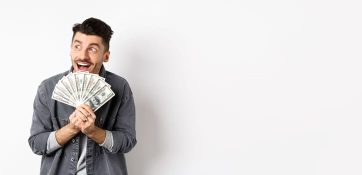 Rich happy man dreaming of shopping wish dollar bills in hands, looking aside pensive and holding money, standing against white background.