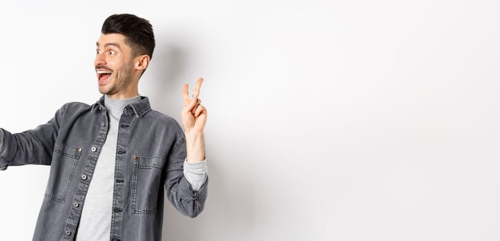 Profile of cheerful young guy taking selfie on smartphone, showing v-sign while make photo on mobile app, standing against white background.