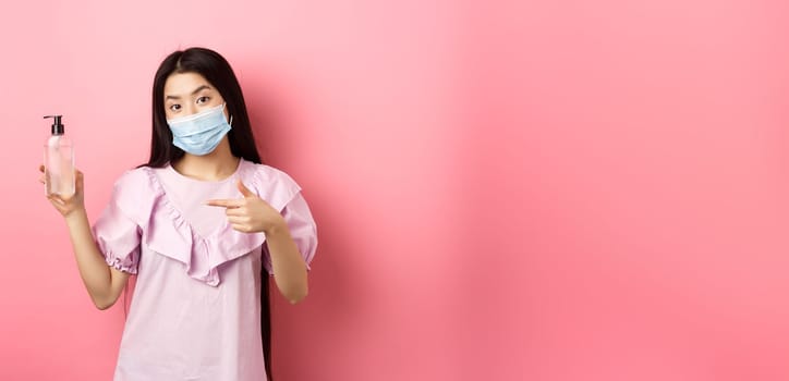 Healthy people and covid-19 pandemic concept. Stylish japanese girl in medical mask showing bottle of hand sanitizer, pointing at antiseptic, standing against pink background.