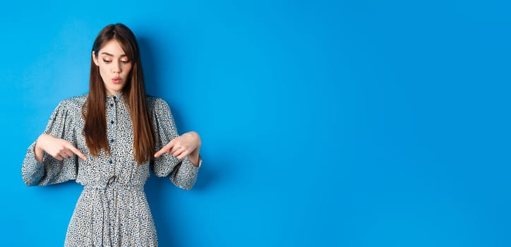 Intrigued beautiful girl in dress say wow, looking and pointing down at advertisement, showing promo with interested face, blue background.