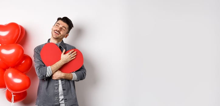 Romantic man hugging big red heart cutout and looking away dreamy, thinking of girlfriend and valentines day, imaging lover, standing on white background.