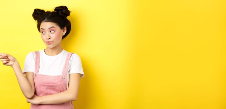 Portrait of stylish asian woman with makeup and summer clothes, pointing and looking left at logo, standing intrigued on yellow background.