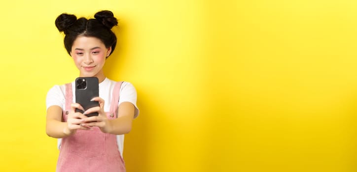 Stylish asian girl taking photo on smartphone, making video with cellphone and smiling, standing on yellow background.