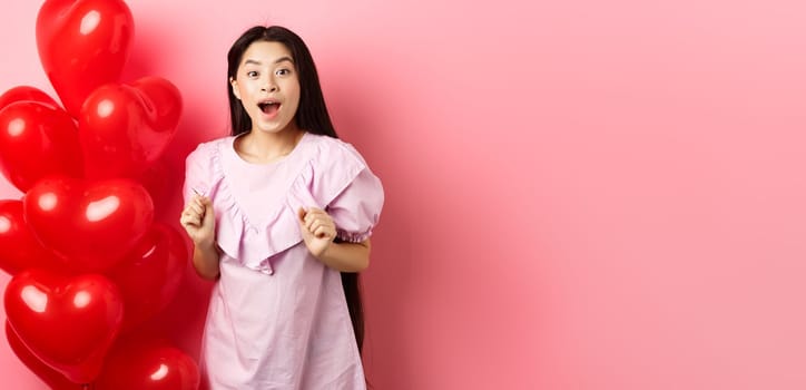 Excited and surprised teenage korean girl open mouth amazed, receive surprise gift on valentines day, looking wondered, standing near heart balloons, pink background.