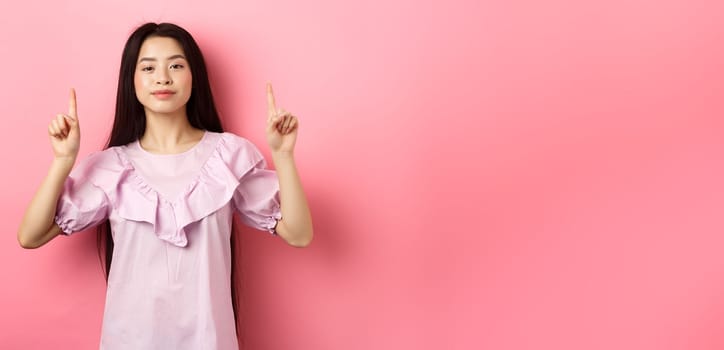 Smiling teen girl asian pointing fingers up at empty space, advertising on pink background.