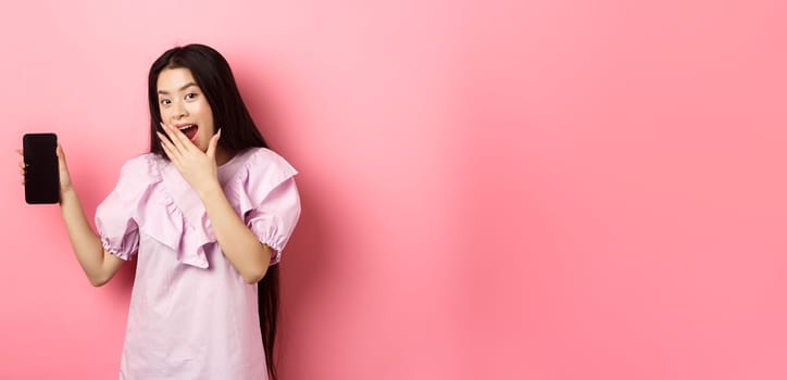 Cheerful asian teen girl showing empty phone screen, laughing and covering mouth with hand, standing in dress against pink background.