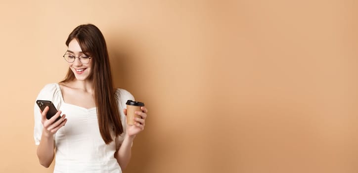 Happy girl chatting on phone and drinking coffee, wearing eyewear and white blouse, smiling at smartphone screen, beige background.