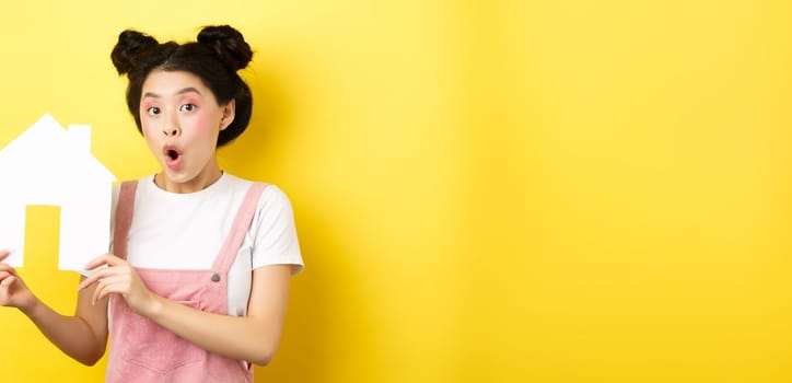 Real estate and family concept. Excited asian teen girl showing paper house cutout and gasping amazed, standing on yellow background.