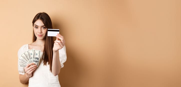 Young woman showing plastic credit card, prefer contactless payment instead of dollar bills, standing on beige background.