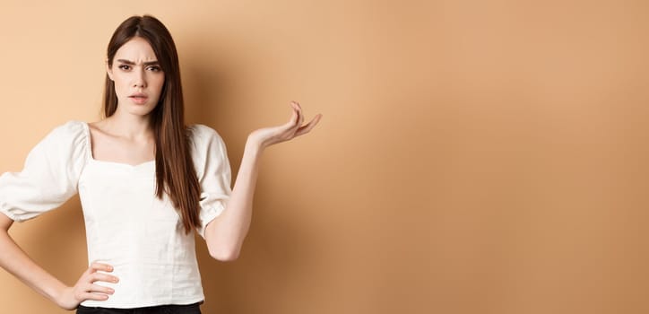 So what. Confused and annoyed young woman raising hand up and staring bothered at camera, cant understand wtf going on, standing on beige background.
