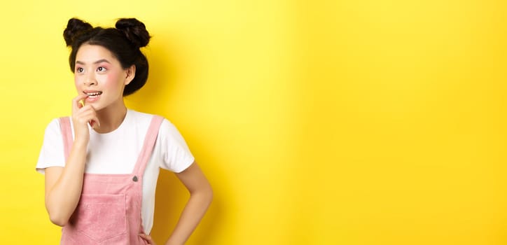 Pensive asian girl feeling excited, biting fingernail and looking left thoughtful, making choice, standing in summer clothes against pink background.