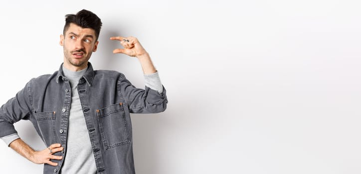 Pensive young man looking aside and thinking, showing small size object, pondering something little or tiny, standing on white background.