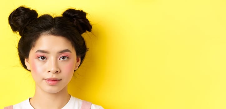 Head portrait of stylish asian girl with bright makeup and hairbuns, looking at camera, standing on yellow background.