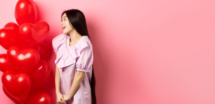 Romance and love concept. Romantic asian girl looking with affection and sympathy at empty space, standing near red hearts balloons from lover, pink background.