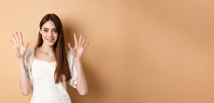 Attractive young woman show fingers number ten, smiling and looking confident, standing on beige background.