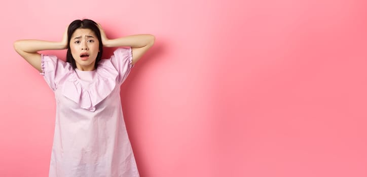 Frustrated asian teen girl panicking, holding hands on head and screaming scared, standing anxious on pink background.