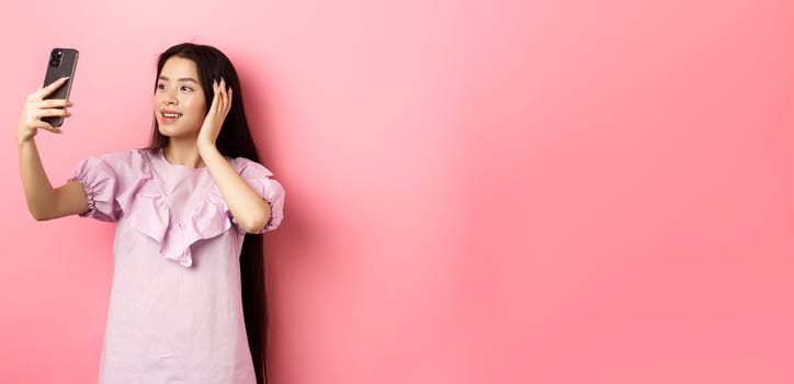 Stylish asian girl blogger taking selfie on mobile phone, posing for smartphone photo, standing in dress against pink background.