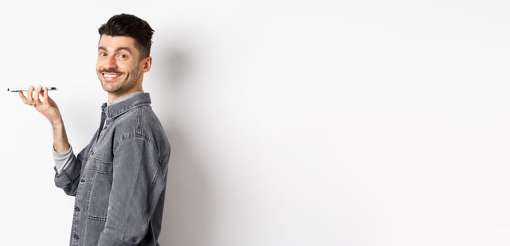 Profile portrait of young man smiling at camera, recording voice to translate word or leave message, talking on speakerphone, standing against white background.