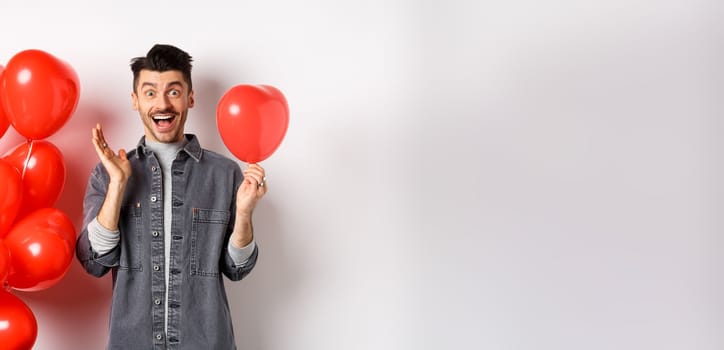 Valentines day concept. Cheerful young man celebrating love holiday, standing near red heart balloons and looking surprised, scream of joy, white background.