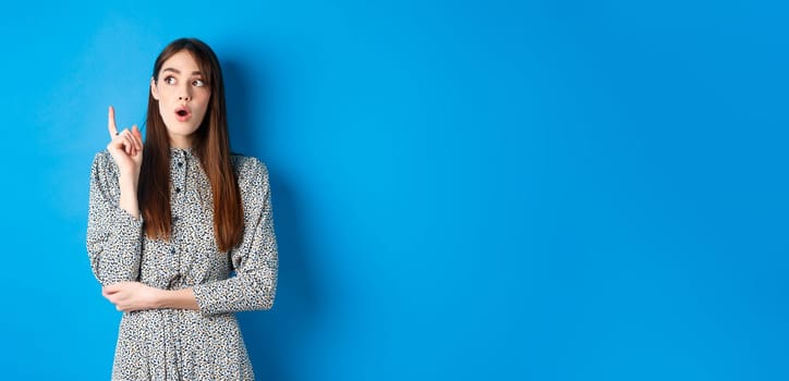 Thoughtful attractive girl in dress pitching an idea, raising finger and looking up, have a plan, standing on blue background.