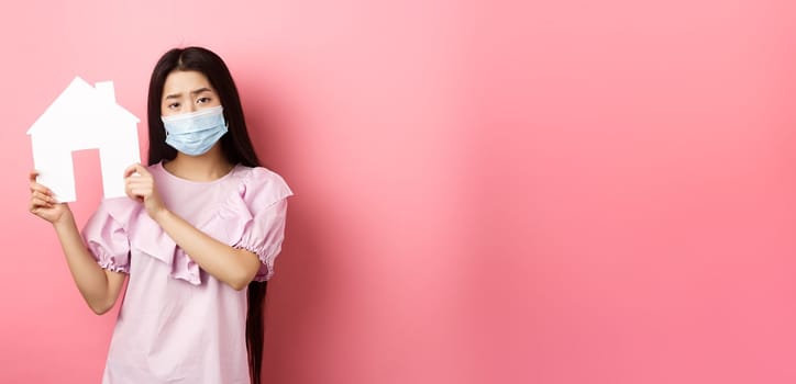 Real estate and pandemic concept. Gloomy girl showing paper house cutout, wearing medical mask and dress, standing against pink background.