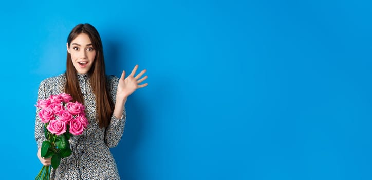 Valentines day concept. Surprised pretty girl holding bouquet of flowers from lover, looking amazed and happy at camera, standing on blue background.