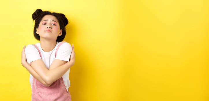 Lonely sad teen girl hugging herself, wanting to relationship and cuddles, standing alone on yellow background.