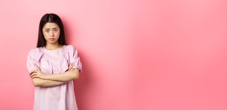 Silly asian girl feel unfair, cross arms on chest and sulking, frowning at camera, standing offended against pink background.
