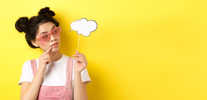 Summer and fashion concept. Stylish glamour asian girl in sunglasses, holding comment cloud party mask and looking aside, yellow background.