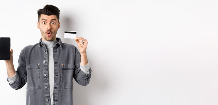 Excited handsome man showing plastic credit card and mobile phone screen, standing on white background.