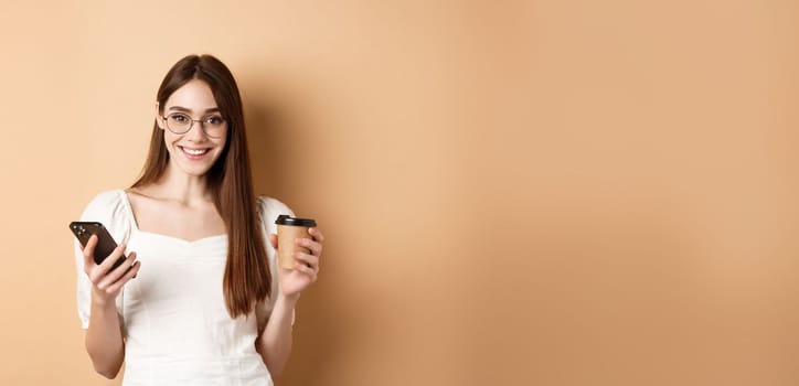 Happy caucasian woman in glasses standing with cup of coffee and smatphone, standing on beige background. Copy space