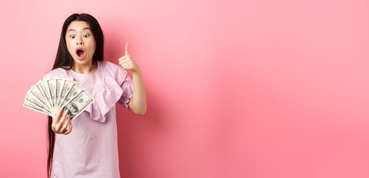 Excited teen girl holding big amount of money, showing dollar bills and thumbs up, standing amazed on pink background.