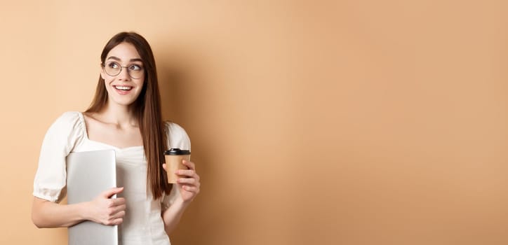 Happy young woman drinking coffee and holding laptop, going study, looking aside with cheerful smile, standing on beige background.