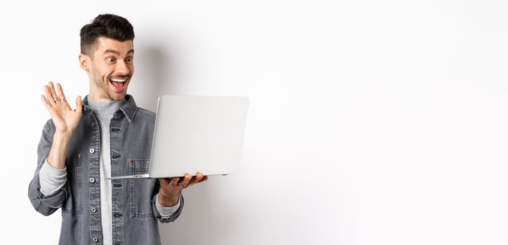 Excited man waiving hand at laptop, video chatting on computer and smiling friendly, standing on white background.