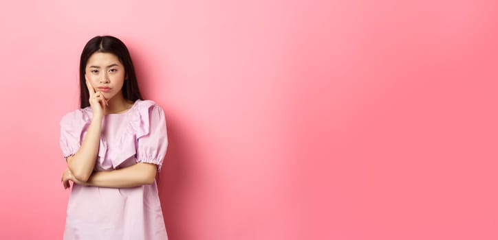 Bored asian teen girl look indifferent at camera, lean face on hand in skeptical pose, standing reluctant against pink background.