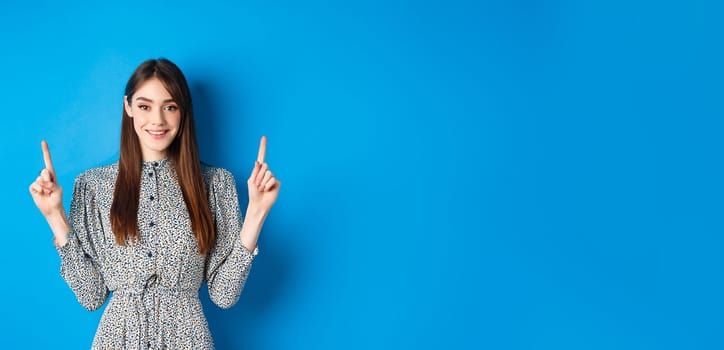 Cute young woman with long hair and vintage dress, smiling and pointing fingers up, advertising on blue background.