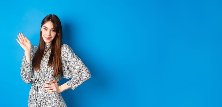 Beautiful lady in dress waving hand and say hello, greeting you with cute smile, standing on blue background.