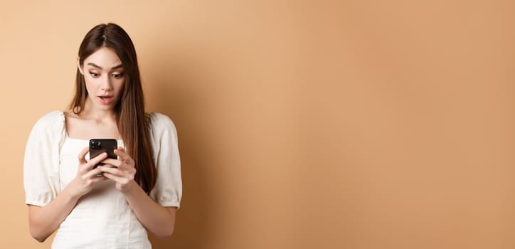 Woman look surprised at smartphone screen, reading shocking message on cell phone, standing amazed on beige background.