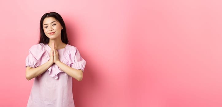 Cute asian girl say thank you, smiling and looking happy, showing namaste gesture in gratitude, standing in dress against pink background.