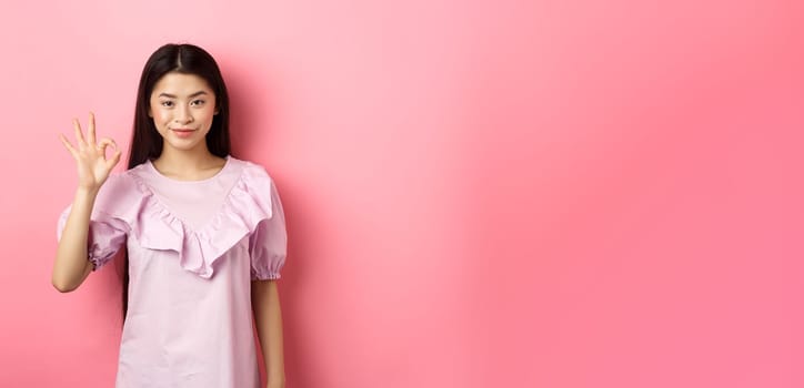 Smiling asian woman in dress showing okay sign, praise and compliment good thing, approve excellent choice, standing on pink background.