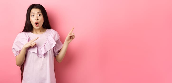 Excited asian woman look amazed, pointing right and gasping fascinated, checking out advertisement, standing on pink background.