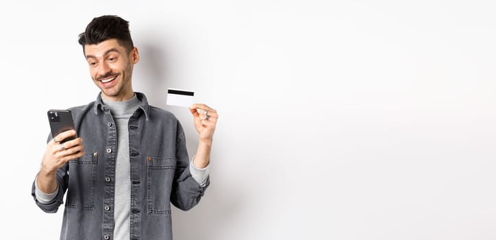 Online shopping concept. Handsome man buying in internet, showing plastic credit card and look at smartphone, standing against white background.
