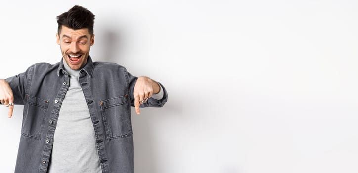 Excited european man looking and pointing fingers down, looking happy at advertisement, standing in denim jacket on white background.